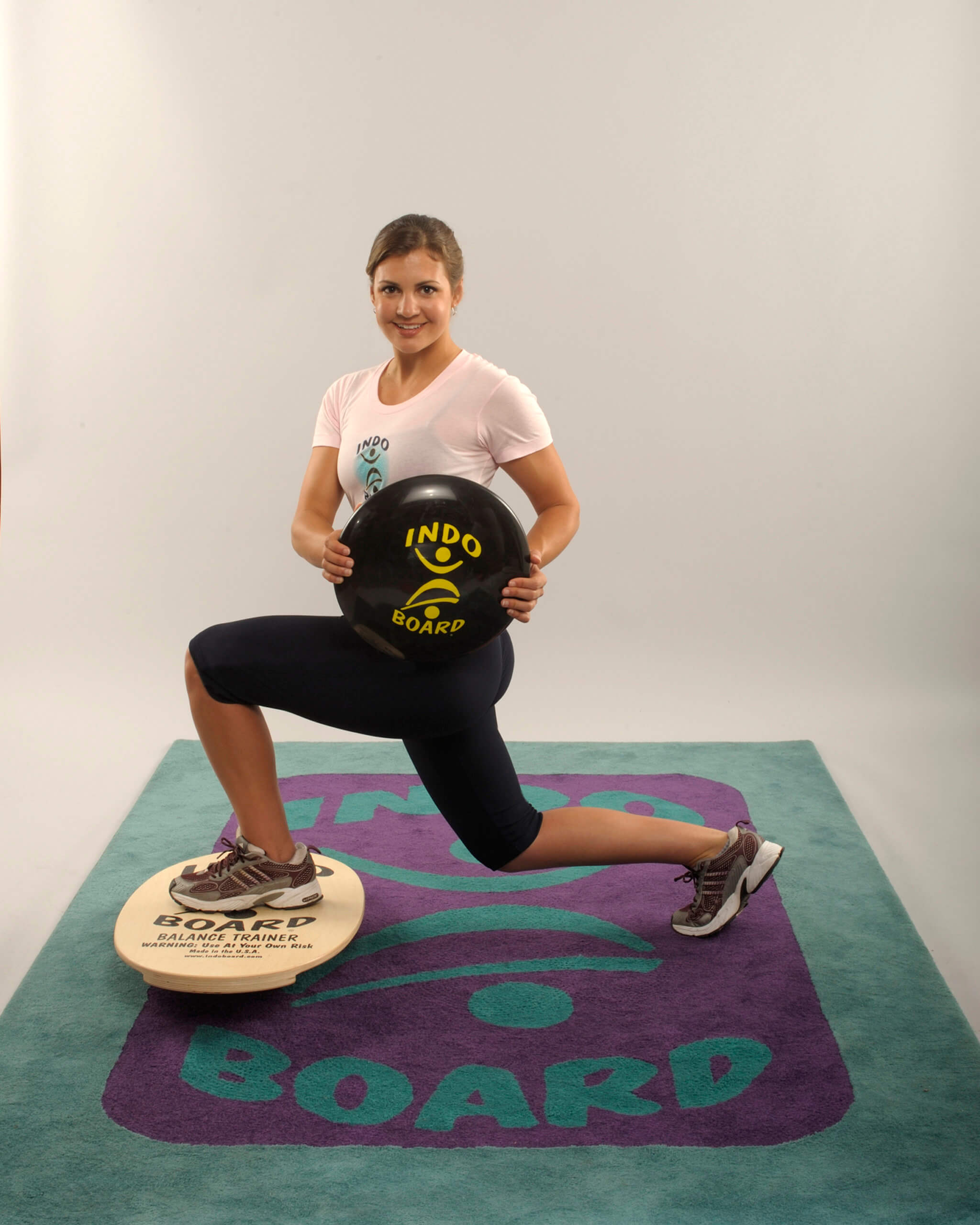 Woman with left leg bent and placed on a balance board, and her rigth leg extended and bent behind her, doing a lunge with the front leg balanced on the Indo Board instead of the floor.