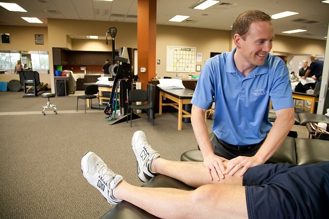 A male physical therapist treats another man's knee at a PT clinic.