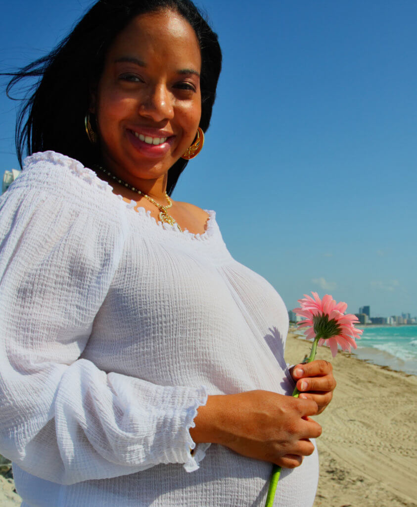 Pregnant Black woman, standing on a beach by the ocean and holding a pink flower.
